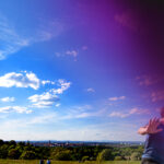 Alttext: Weite Landschaft unter einem geteilten Himmel: links leuchtend blau, rechts dunkel violett-schwarz verfremdet; eine Stadt liegt am Horizont, rechts läuft ein Mensch mit erhobener Hand aus dem Bild.